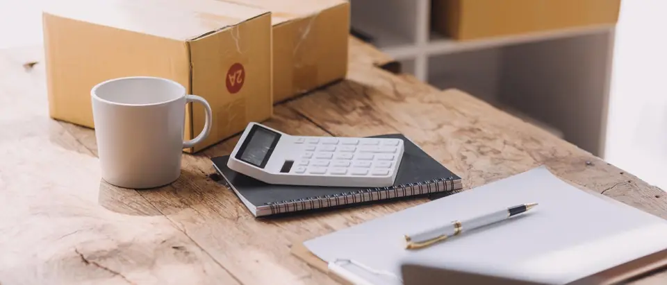 A calculator on a notebook, a clipboard with a pen and a mug beside taped cardboard boxes on a wooden table