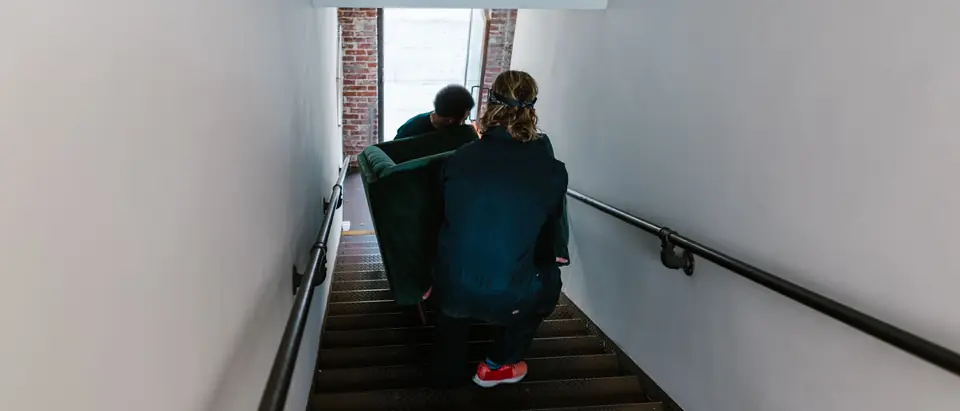 Two movers seen from behind carry a green sofa down a narrow stairway toward an open street door