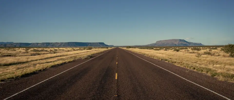 A straight two-lane highway running to the horizon through dry West Texas grassland, with flat-topped mesas in the distance