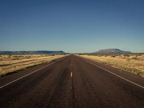 A straight two-lane highway running to the horizon through dry West Texas grassland, with flat-topped mesas in the distance