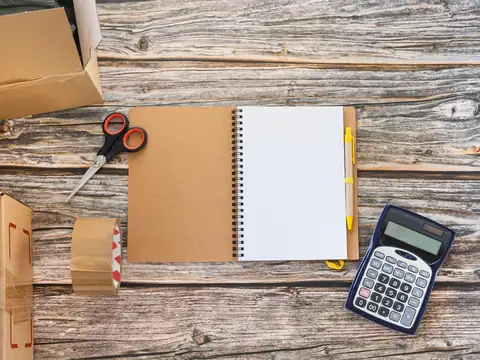 An open blank notepad with a pen, a calculator, scissors and a roll of packing tape among cardboard boxes on a wooden table