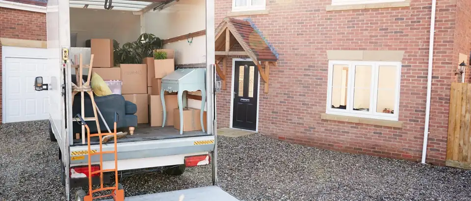 A box truck with its tail lift lowered and a hand truck standing on it, loaded with cartons and furniture, parked on the gravel drive of a red brick house