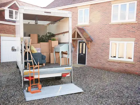 A box truck with its tail lift lowered and a hand truck standing on it, loaded with cartons and furniture, parked on the gravel drive of a red brick house
