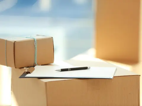 A clipboard with a ruled form and a pen resting on a cardboard box, beside a smaller box tied with string
