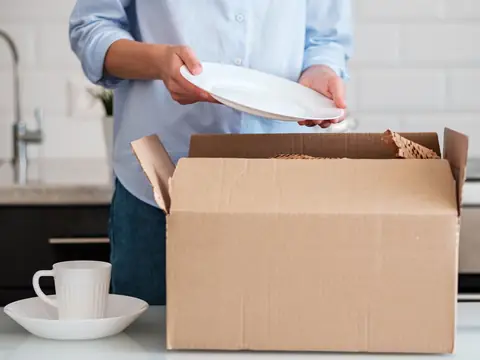 Hands holding a white plate over an open carton lined with packing paper on a kitchen counter, with a cup and saucer beside it