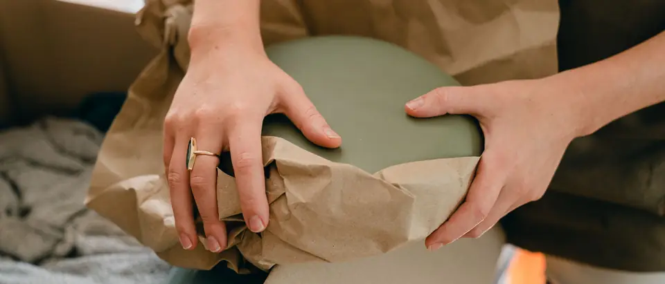Two hands folding brown packing paper around a green ceramic dish above an open moving box