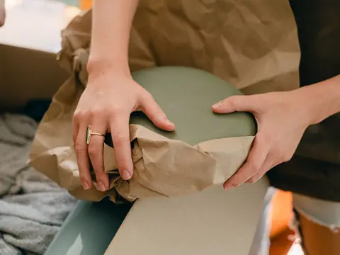 Two hands folding brown packing paper around a green ceramic dish above an open moving box